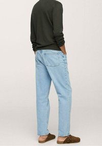 Personne portant un jean bleu clair, un haut à manches longues foncé et des chaussures marron à enfiler, debout, les mains dans les poches, vue de dos.