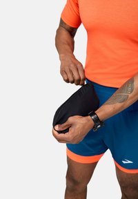 Črn žep z zadrgo, ki ga drži desna roka, oblečen v oranžno majico s kratkimi rokavi in modre kratke hlače z oranžnimi poudarki. Vidna je ura na zapestju.