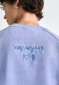 Blå hettegenser med kontrastsøm og brodert tekst, med "MILANO EST" og "1978" i en glansfull, dryppende effekt.