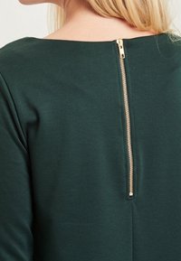 Femme portant un haut vert foncé avec une fermeture éclair dorée courant verticalement le long du haut du dos, cheveux blonds partiellement visibles au niveau du cou.