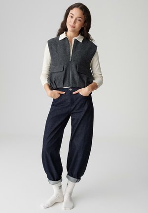 Gray wool vest with zip closure, two front pockets, worn over a cream long-sleeve shirt, paired with dark blue rolled-up jeans and white socks.