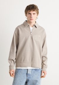 Hanorac bej cu fermoar, cu guler, logo brodat "CK", textură ribă, și mâneci lungi. Purtat peste un tricou alb. Blugi de culoare bleu deschis.