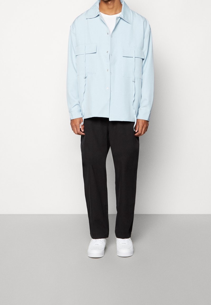 Ljust blå långärmad skjorta med två bröstfickor, knappstängning framtill, i kombination med svarta löst sittande byxor och vita sneakers.