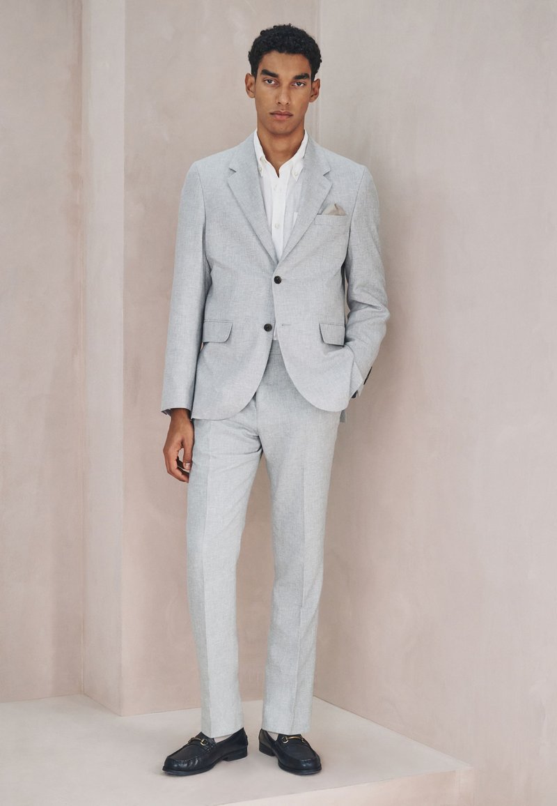 Un homme se tient debout, une main dans la poche, portant un costume gris clair, une chemise blanche et des chaussures habillées noires sur un fond beige uni.