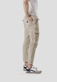 Pantalones cargo beige con múltiples bolsillos, cintura ajustable y puños elásticos. Combinados con zapatillas deportivas blancas, grises y negras.