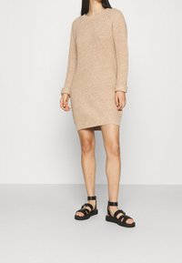 Robe-pull beige tricotée avec manches longues et poignets côtelés, portée avec des sandales noires à lanières. Texture lisse et coupe décontractée.