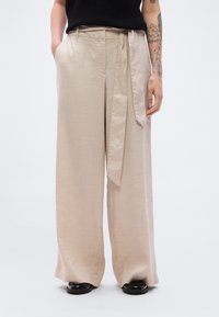 Μπεζ παντ ελόνι λινό με φαρδιά πόδια και ζώνη στη μέση για δέσιμο, με πλαϊνές τσέπες και λεία υφή· φοριέται με μαύρα παπούτσια.