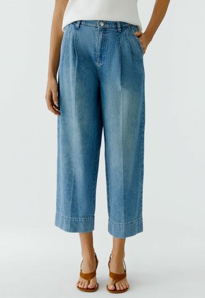 Vrouw draagt wijde cropped blauwe spijkerbroek met opgerolde zoom, voorplooien en bruine sandalen met open neus, staand met één hand in de zak.