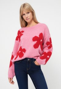 Jovem mulher vestindo um suéter de malha rosa com grandes flores vermelhas e jeans escuros, posando com uma mão no bolso contra um fundo branco.