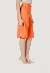 Culotte arancioni a vita alta realizzati in tessuto leggero, caratterizzati da pieghe e una texture liscia, abbinati a scarpe con tacco nude.