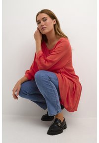 Žena čuči uz bijeli zid, obučena u labavu crvenu bluzu, plave traperice i crne mokasine sa zlatnim detaljima.