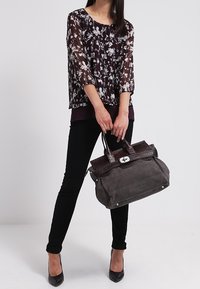 Durchsichtiger schwarzer Blumenblusen, dunkelgraue Jeans und schwarze High Heels. Hält eine zweifarbige grau-braune Handtasche mit strukturiertem Design und silbernem Zubehör.