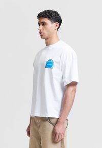 Άνδρας που φοράει λευκό oversized T-shirt με μπλε κηλίδα με την επιγραφή «ETERNITY» στο στήθος και μπεζ παντελόνι με κολλημένη ασημένια αλυσίδα.