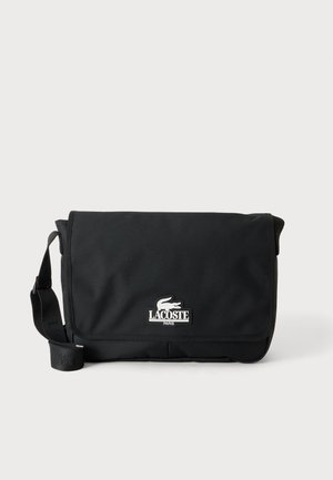 Sac messager noir Lacoste avec rabat avant et bandoulière réglable, avec logo crocodile blanc et texte "Lacoste Paris".