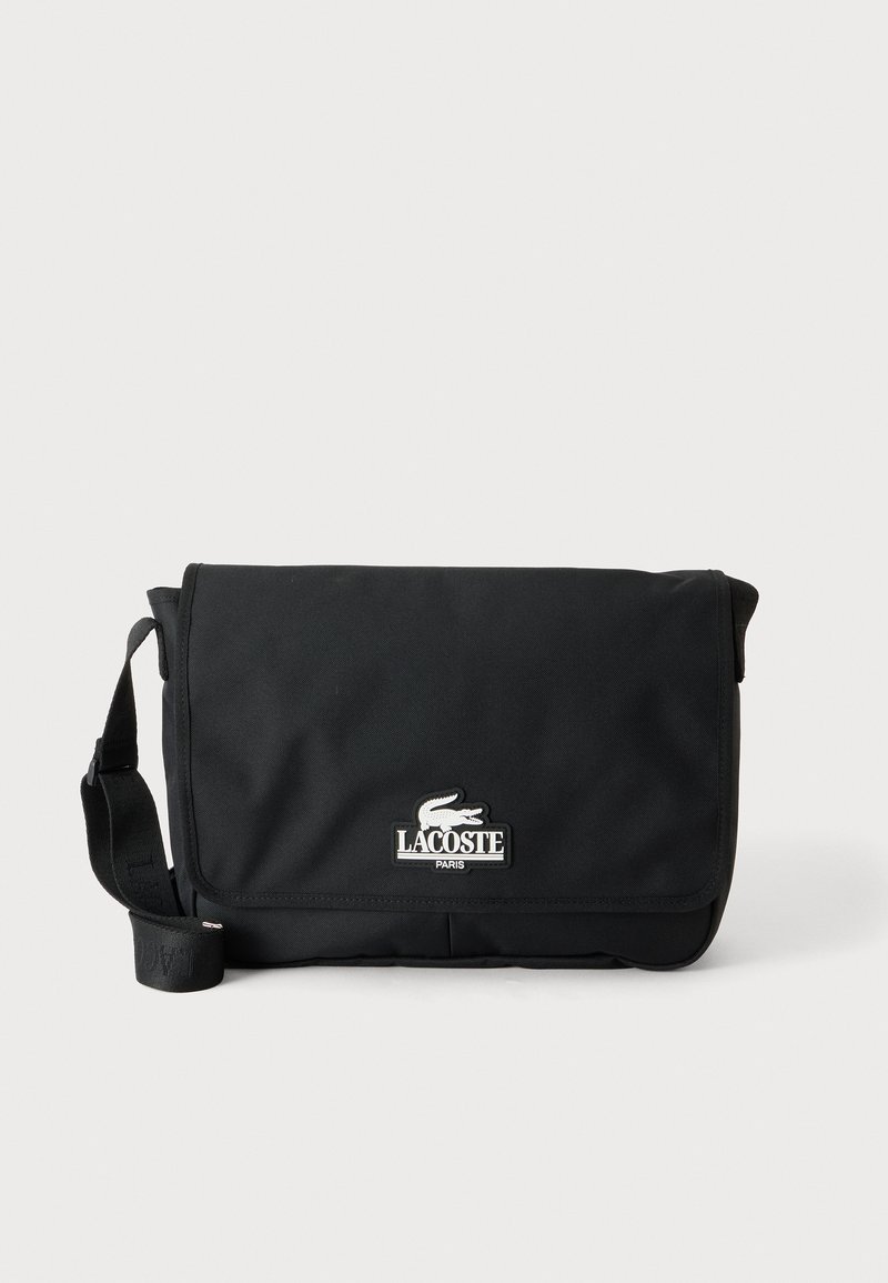 Черна чанта за през рамо Lacoste с преден капак и регулируема презрамка, с бяло лого на крокодил и текст "Lacoste Paris".