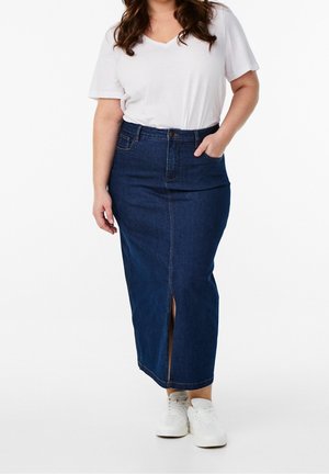 Kvinde iført hvid V-hals kortærmet skjorte, mørkeblåt denim midinederdel med frontslids og hvide sneakers, stående med den ene hånd i lommen.