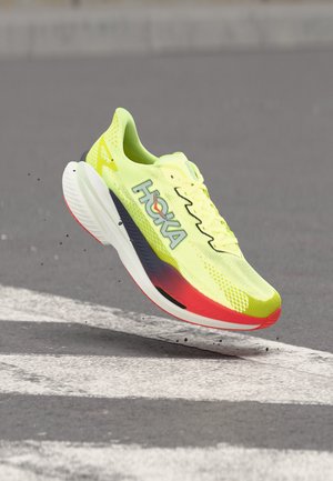 Scarpa da corsa giallo brillante e rossa con marchio HOKA nell'aria sopra una strada grigia con segnaletica bianca.