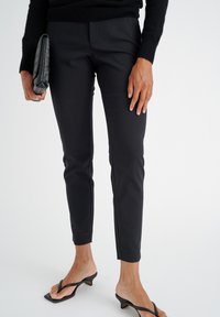 Černé kalhoty na míru se slim fit, mírně zkrácené nad kotníky, spárované s černými sandály na podpatku a texturovanou kabelkou na clutch.