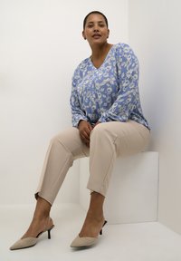 Himmelbluse mit langem Arm und floralem Muster, beigefarbene zugeschnittene Hose und beigefarbene spitze Slingback-Absätze, sitzend auf einem weißen Würfel in einem minimalistischen Ambiente.