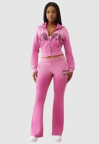 Różowy dres dwuczęściowy składający się ze skróconej bluzy na zamek z nadrukiem oraz dopasowanych spodni o rozszerzanych nogawkach. Wykonany z miękkiej, teksturowanej tkaniny.