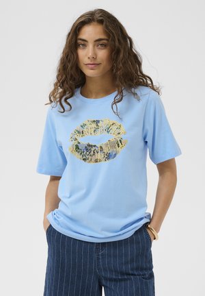 Jeune femme aux cheveux bouclés portant un t-shirt bleu clair avec un grand imprimé floral en forme de lèvres, un pantalon à fines rayures foncées et un bracelet, mains dans les poches.