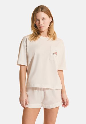 Jeune femme aux cheveux châtain clair portant un haut crème à manches courtes avec une petite broderie florale sur la poche poitrine et un short assorti.