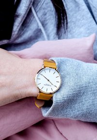 Reloj de oro con una esfera blanca y marcadores negros. Presenta una correa de cuero amarilla. La mano descansa sobre un suéter gris claro contra un fondo rosa.