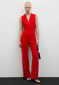 Crvena bezrukavna kombinezon s dubokim V-izrezom, uskim strukom i širokim hlačama. Gladak materijal s minimalnim detaljima. Crna clutch torba i sandale.