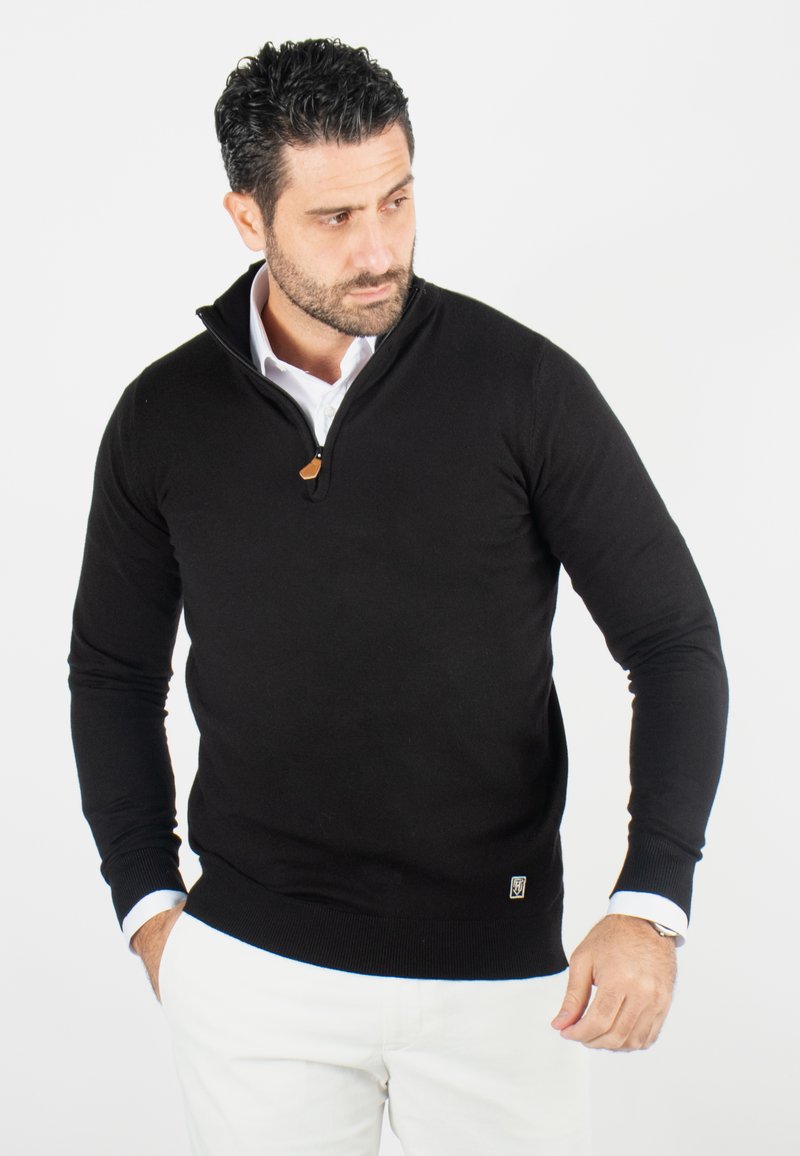 Pull noir zippé avec manches longues, poignets et ourlet côtelés, doté d'un accent en cuir au col. Porté sur une chemise blanche.