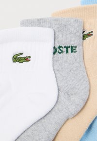 Σετ τριών κάλτσων: λευκή με πράσινο λογότυπο, γκρί με πράσινο κείμενο "LACOSTE" και ανοιχτό μπεζ. Μαλακό ύφασμα, ριμπ μανίκια και διάφορα χρώματα.