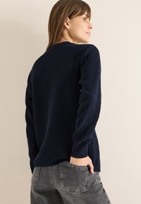 Maglione blu navy a costine con maniche lunghe, scollo tondo e vestibilità morbida, abbinato a jeans di denim grigio con una tasca posteriore.