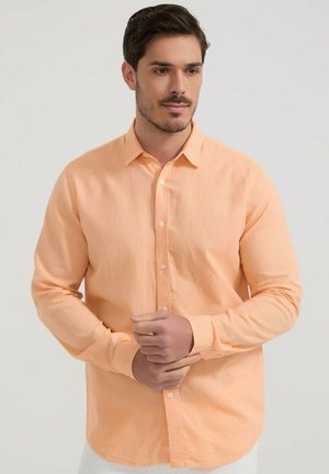 Homme portant une chemise pêche à manches longues boutonnée, debout devant un fond clair uni, ajustant un poignet.