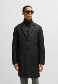 Piumino nero con un design su misura, chiusura a bottoni e tasche laterali. Indossato sopra un capo nero a collo alto, caratterizzato da una texture liscia e lucida.