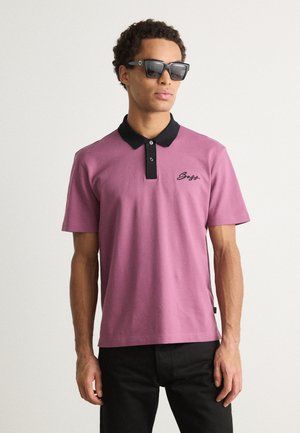 Rosa polo-shirt med sort krave og knapper, med et broderet logo på venstre bryst; parret med sorte solbriller og mørke bukser.