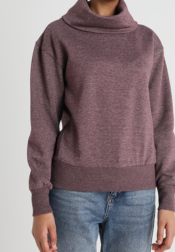 Persona vestida con un suéter de cuello alto de manga larga morado, combinado con jeans azules, mostrada desde el cuello hasta las caderas sobre un fondo gris.