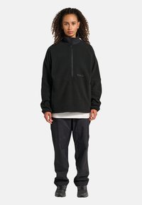 Zwarte fleece pullover met een hoge kraag en een halve ritssluiting, voorzien van een geborduurd "HALO" logo. Gecombineerd met losse zwarte broek en schoenen.