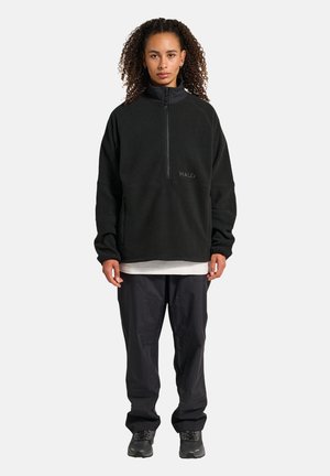 Zwarte fleece pullover met een hoge kraag en een halve ritssluiting, voorzien van een geborduurd "HALO" logo. Gecombineerd met losse zwarte broek en schoenen.