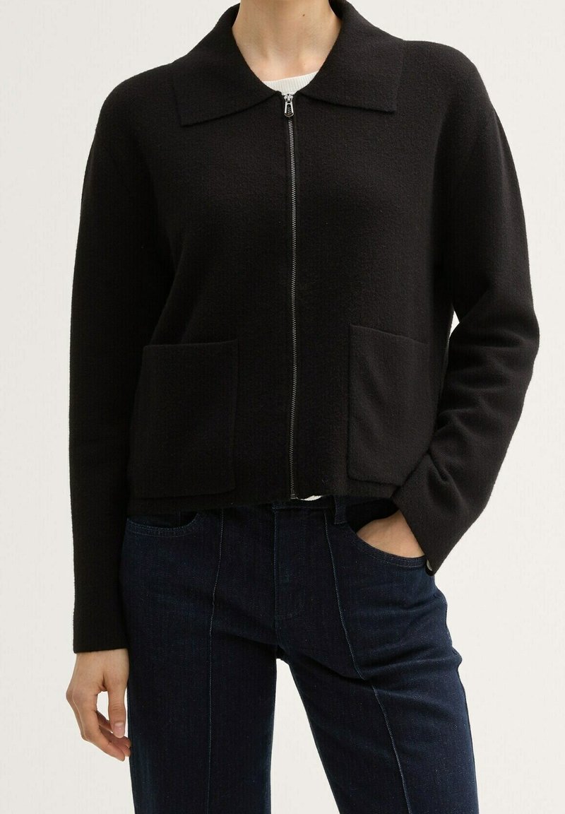 Cardigan noir zippé avec col et deux poches avant, porté sur un jean bleu foncé, main dans la poche.