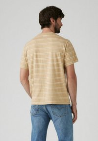 Koszulka z krótkim rękawem w jasnym beżu z cienkimi białymi poziomymi paskami, o luźnym kroju i okrągłym dekolcie, widziana z tyłu.