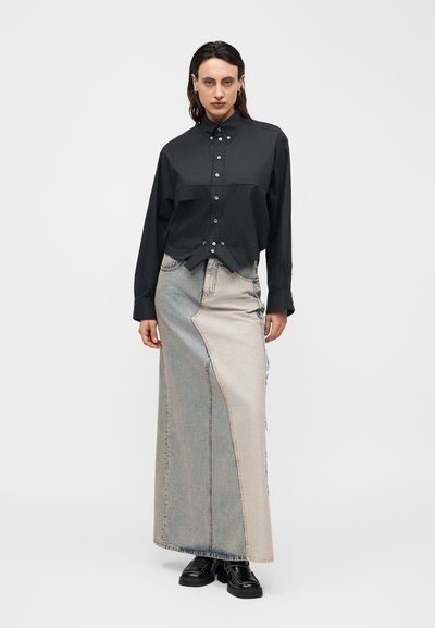 Άτομο που φοράει μαύρο πουκάμισο με κουμπιά και μακριά δίχρωμη φούστα από τζιν με μαύρα παπούτσια, στέκεται μπροστά σε ένα απλό ανοιχτό φόντο.