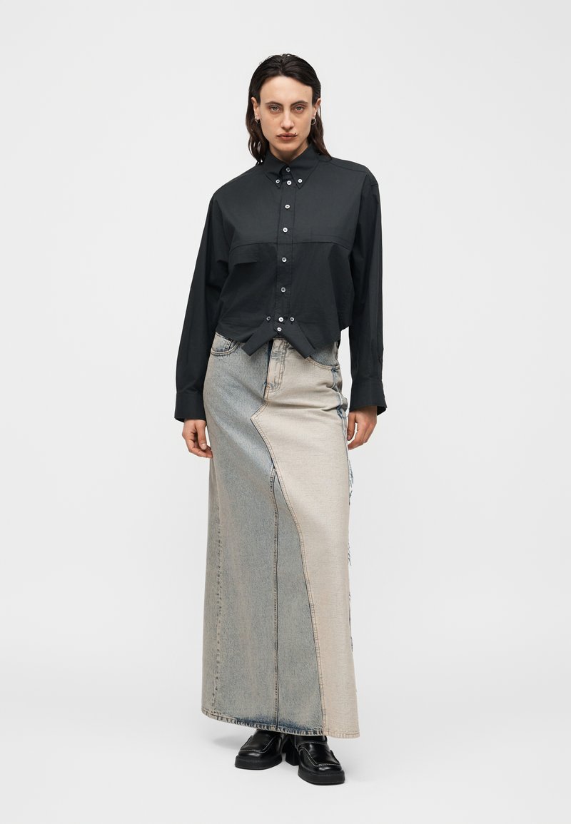 Personne portant une chemise noire à boutons et une longue jupe en denim bicolore avec des chaussures noires, debout devant un fond clair uni.