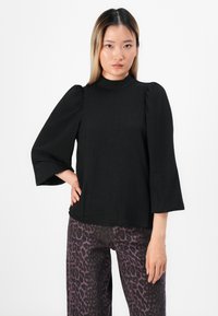 Kvinne med svart puffermet bluse og lilla leopardmønstrede bukser står mot en enkel hvit bakgrunn.