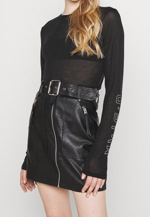 Femme portant un haut noir transparent à manches longues et une jupe en cuir noire taille haute avec une fermeture éclair frontale et une ceinture à boucle.