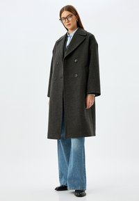 Donkergrijze dubbelzijdige wollen jas met grote revers en knoopdetails, gecombineerd met een lichtblauw shirt en wijde denim jeans.