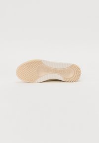 Sohle eines Sneakers mit einer strukturierten, beigen Gummifläche, einem gemusterten Profil und weißen Akzenten. Der Markenname "Scotch & Soda" ist auf der Ferse eingraviert.