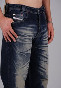 Man wearing faded dark blue jeans with hand resting on left thigh, showing texture and pocket details against a plain background.