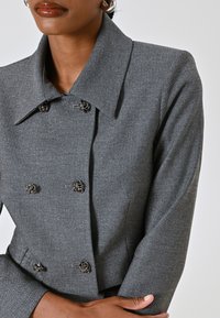 Veste grise à double boutonnage avec un motif en chevrons, dotée d'un grand col et de boutons décoratifs en métal argenté. Texture lisse.