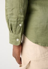 Cămașă din in de culoare verde oliva, cu manșete butonate și detaliu brodat cu logo. Textură fină, hem curbat, purtată peste pantaloni de culoare deschisă.