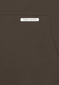 Brauner Sweatshirt-Stoff mit einem kleinen silbernen Metallschild mit der Aufschrift "bazir republiq" in der Nähe der Tasche. Glatte Textur, schlichtes Design.