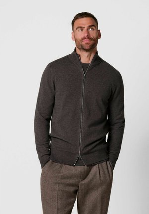 Uomo con la barba che indossa un maglione grigio scuro con zip e pantaloni marroni, in piedi con le mani in tasca davanti a uno sfondo semplice.